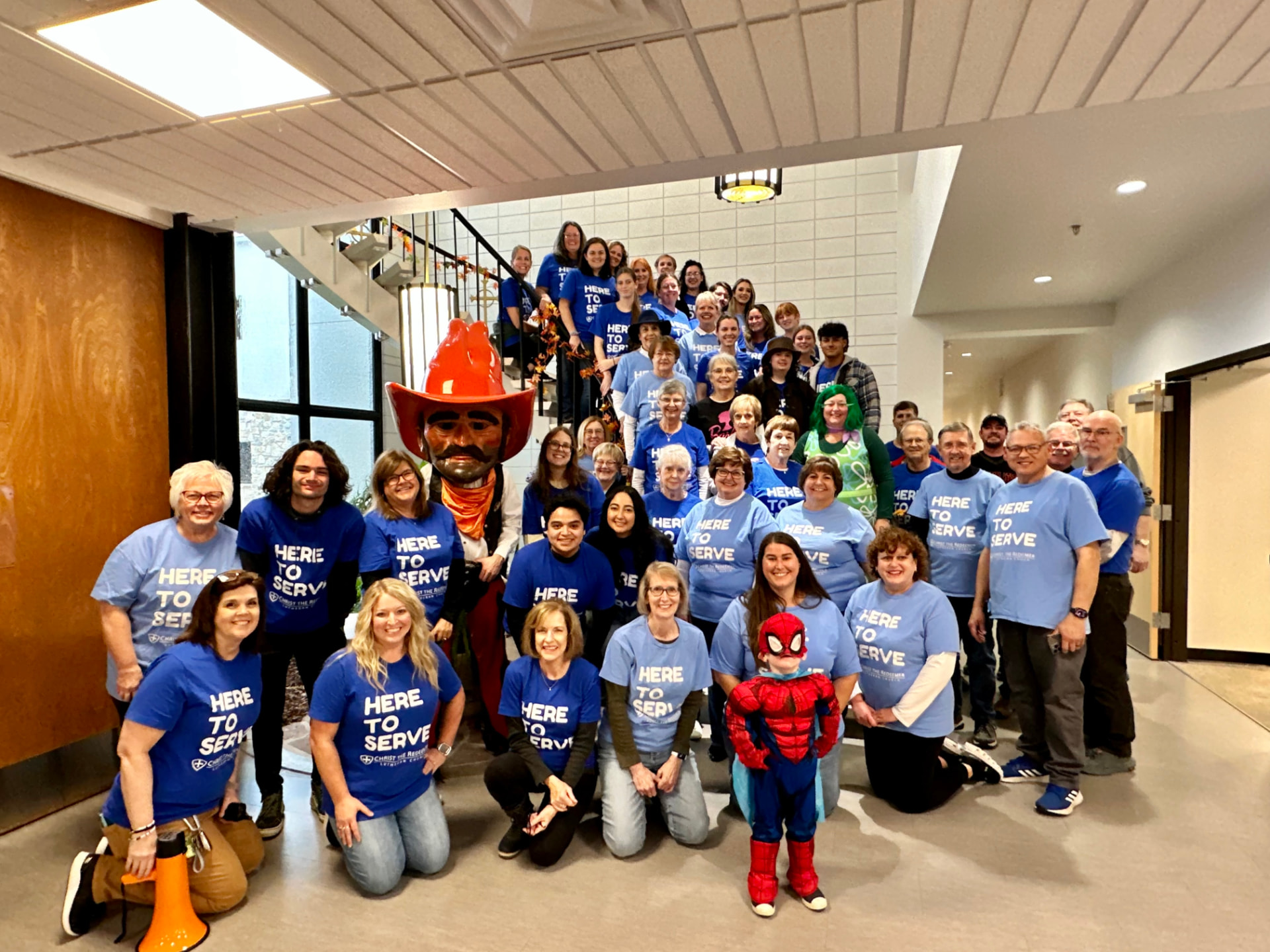 Church fall festival volunteers wearing Here to Serve shirts gathered together indoors